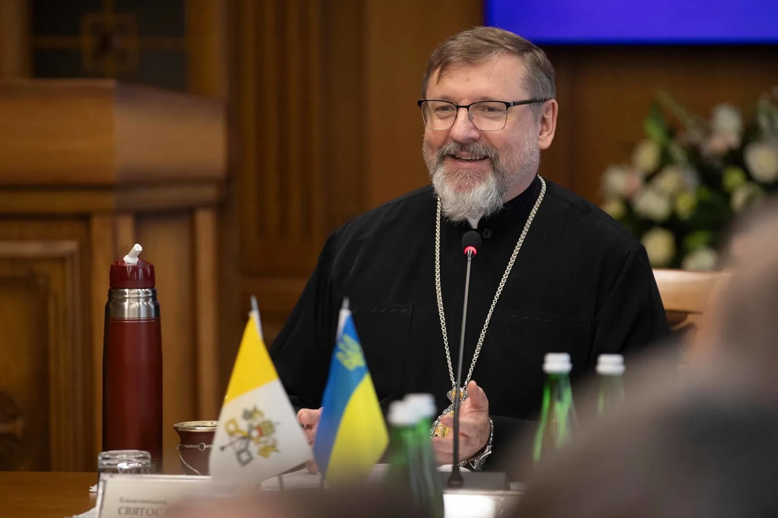 His Beatitude Sviatoslav smiles, wearing black cassock and pectoral cross, with Vatican and Ukraine flags.