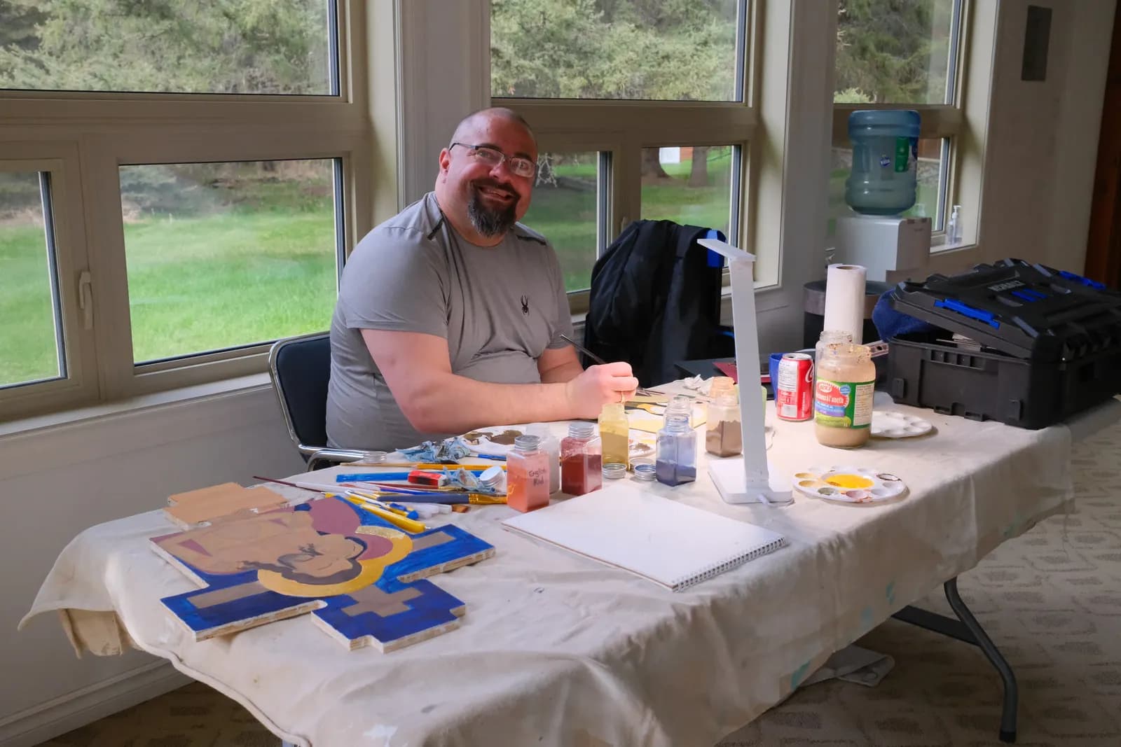 Man smiles while painting an icon of Christ on a cross-shaped wooden panel.