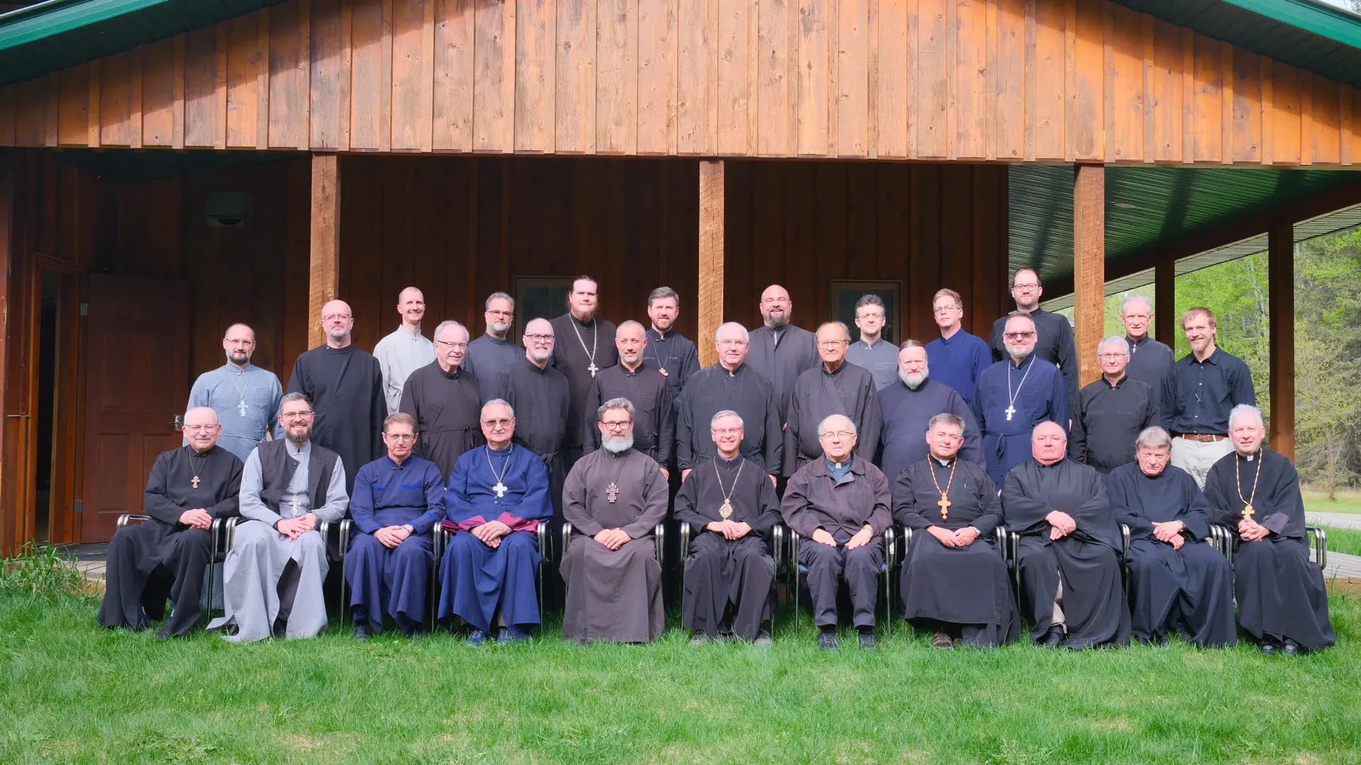 Clergy in cassocks and vestments pose for a group photo outside a wooden building.