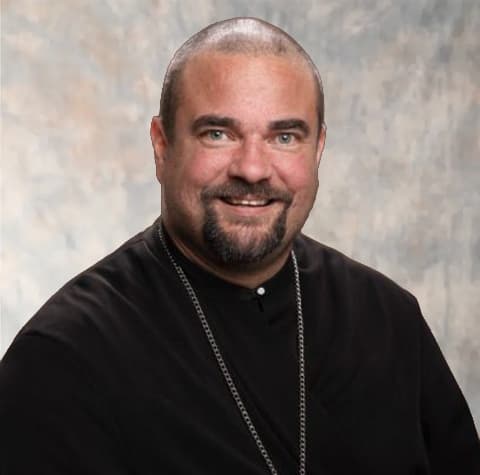 Priest smiles, wearing a black cassock and a silver chain.