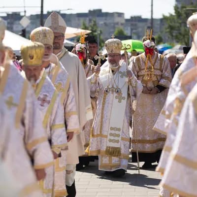 Clergy in ornate vestments and mitres process outdoors, led by a bishop holding a cross.