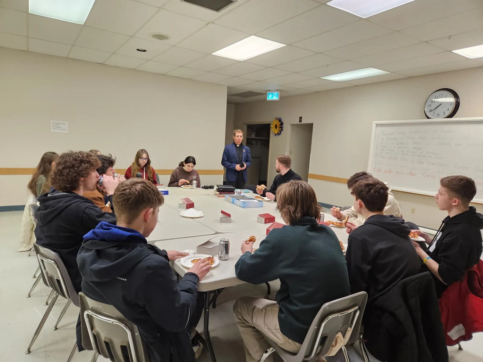 seminarian in clerical attire speaks to a group of young people gathered around a table with pizza.