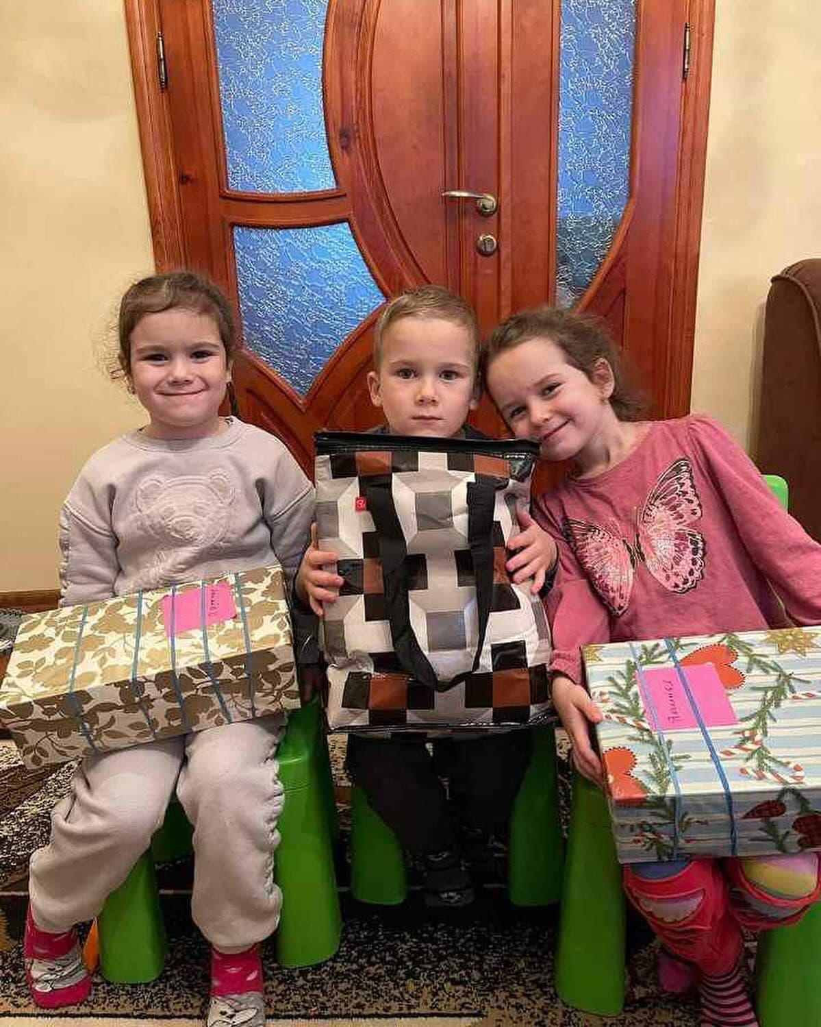Three children sit with gifts in front of a wooden door.