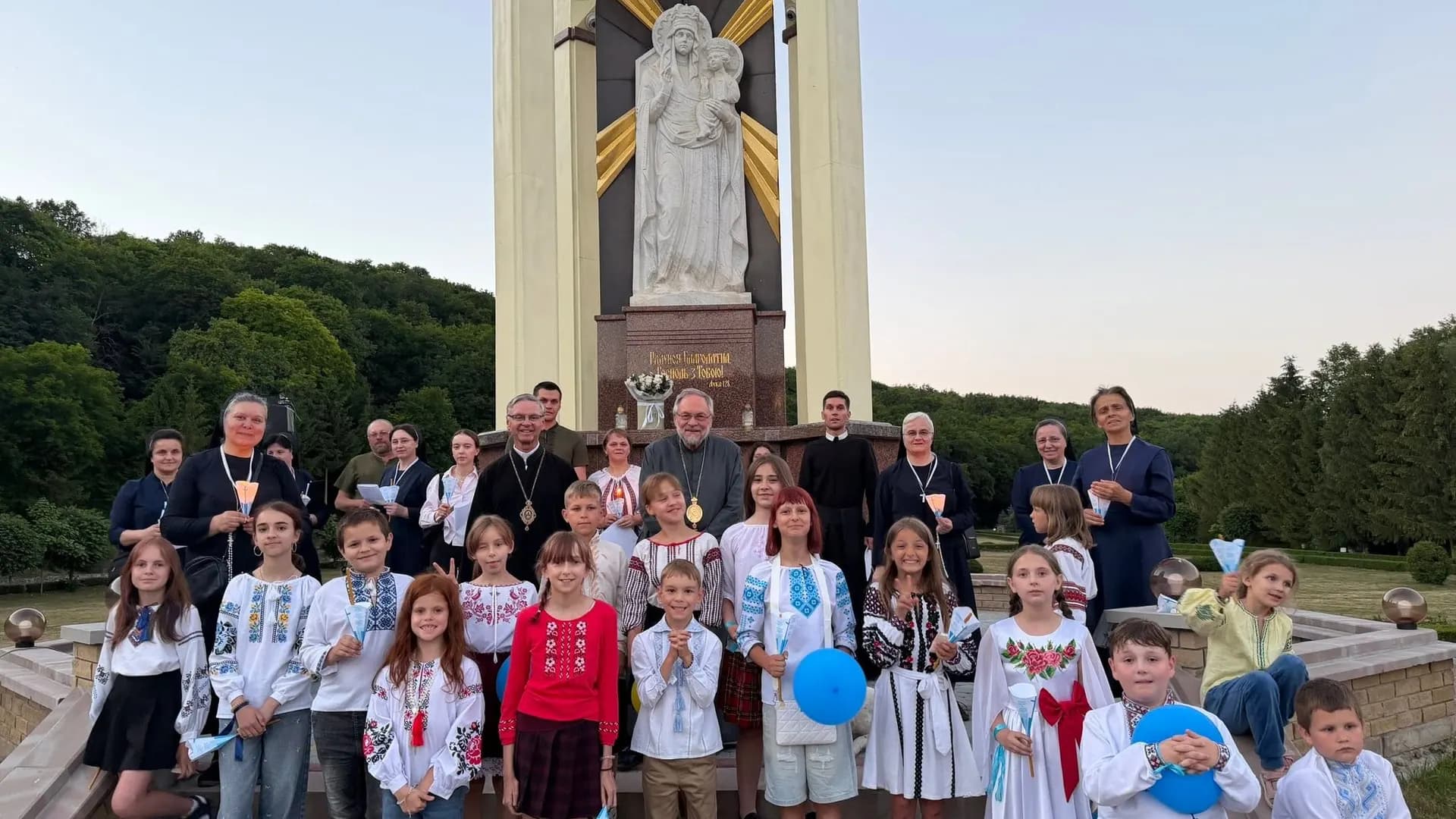 Clergy and children in traditional Ukrainian clothing stand before a statue of the Virgin Mary.