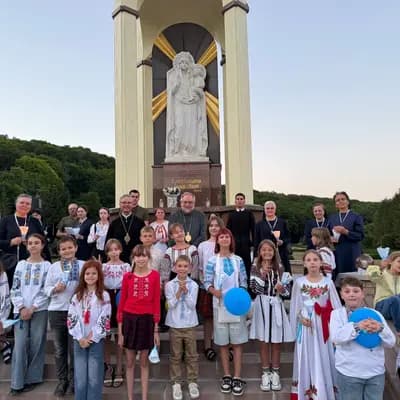 Clergy and children in traditional Ukrainian clothing stand before a statue of the Virgin Mary.