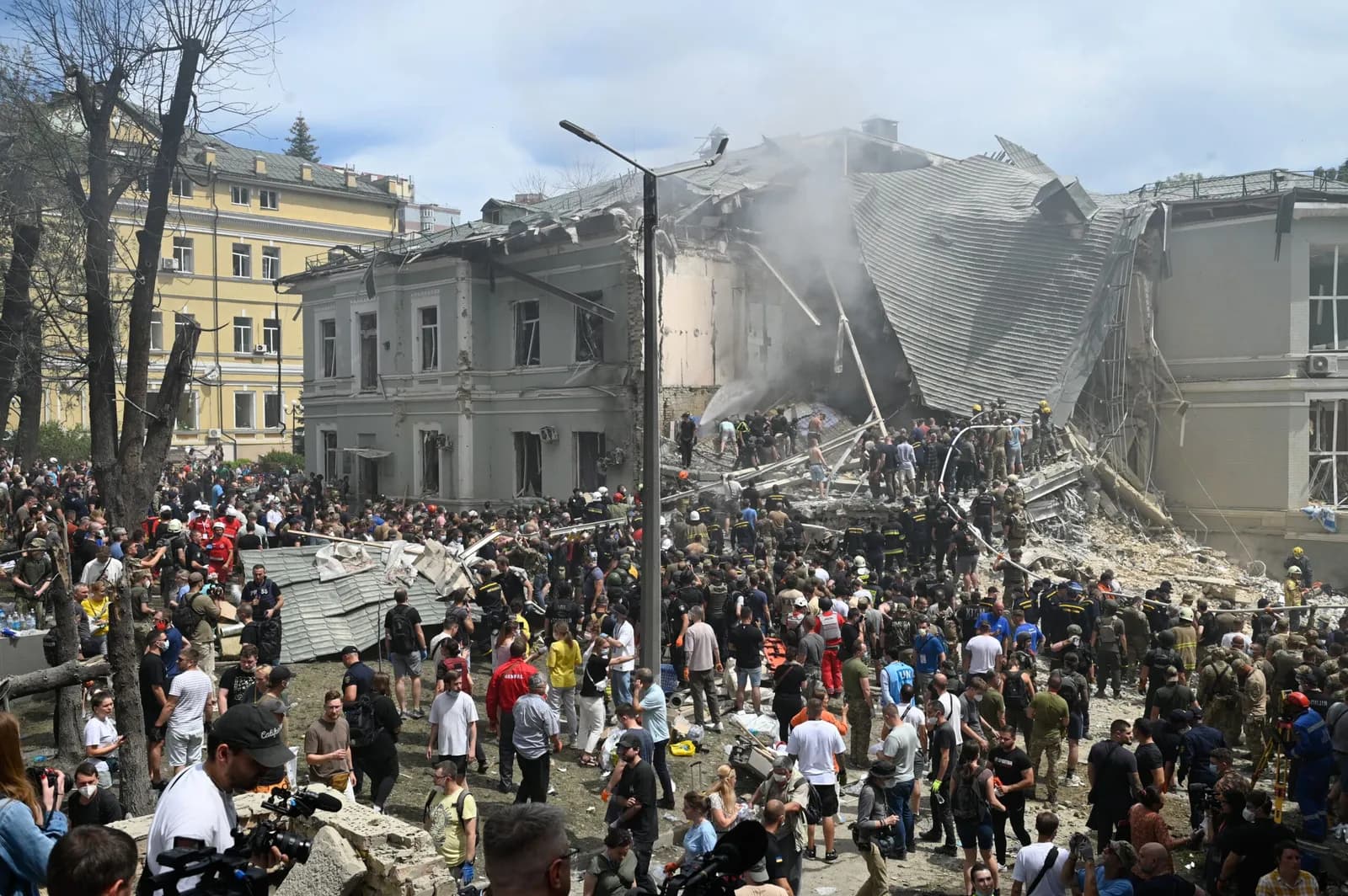 Emergency responders and civilians gather around a damaged building with smoke rising.