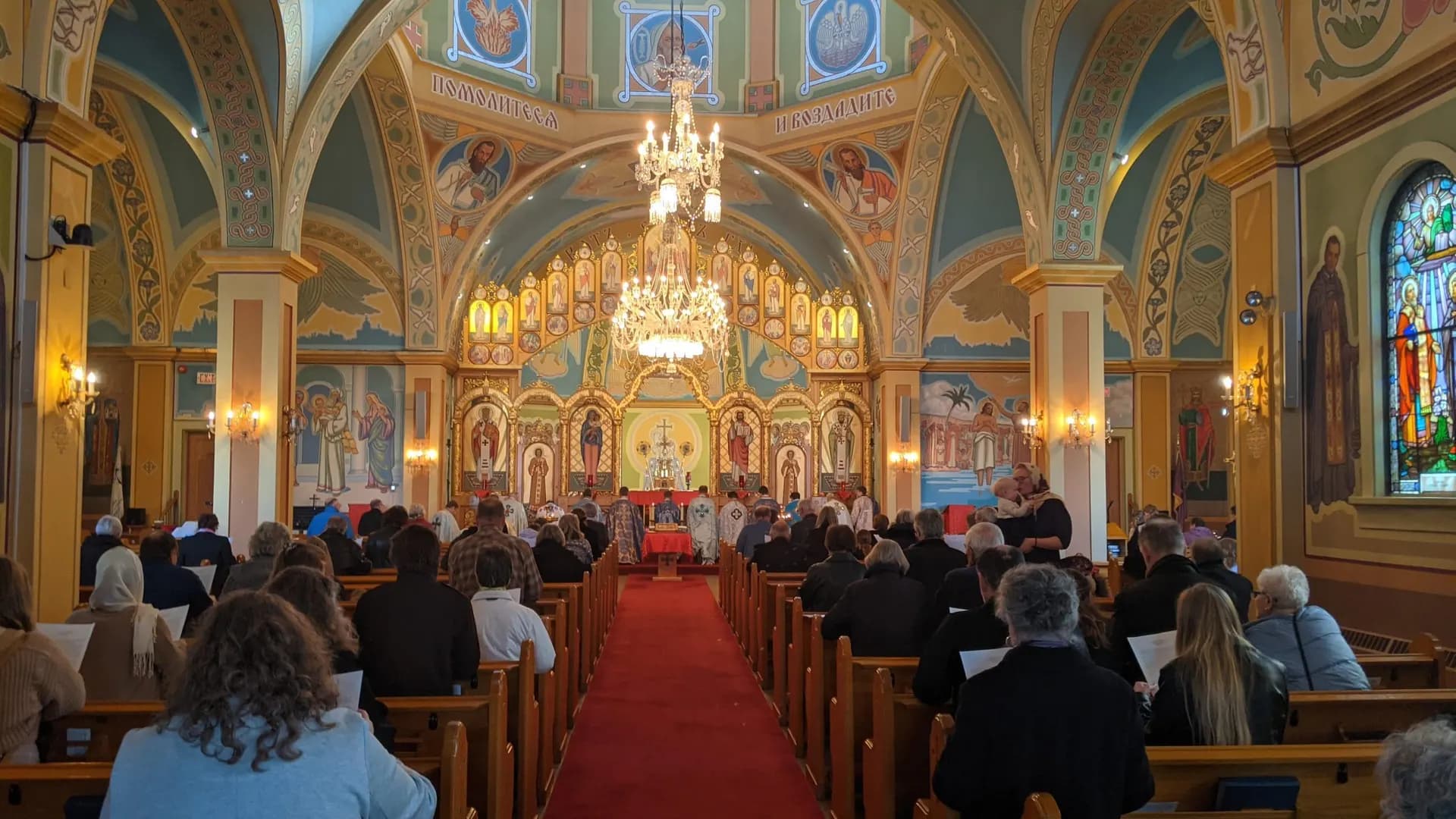 Congregation attends Divine Liturgy in a church with a decorated iconostasis and frescoes.