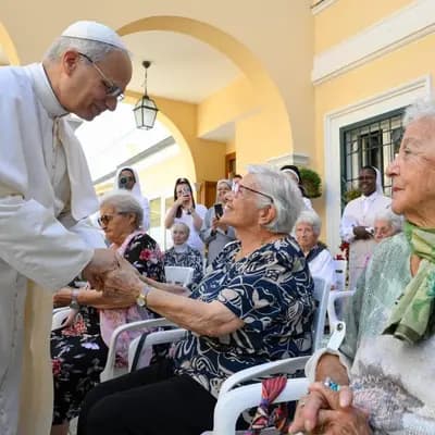 Pope Francis greets elderly women seated outdoors, holding their hands.