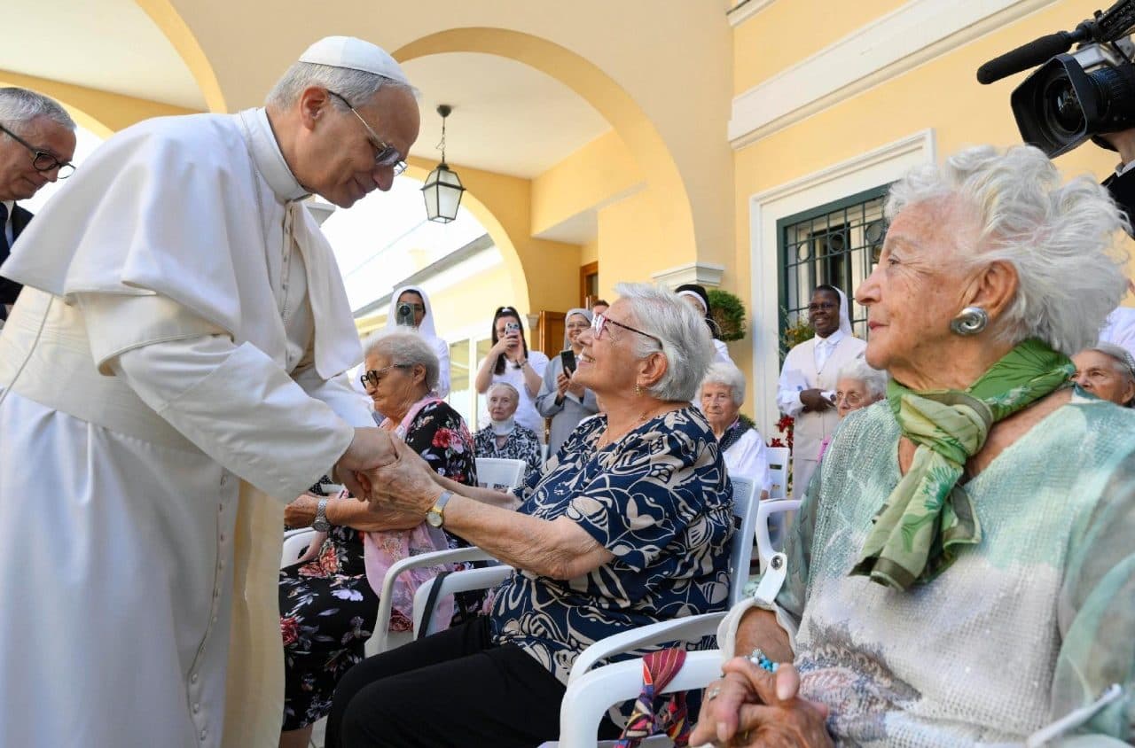 Pope Francis greets elderly women seated outdoors, holding their hands.